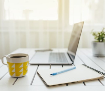a cup of coffee, notepad and pen, and a laptop on a table